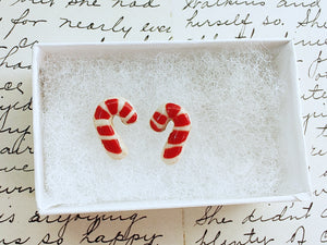 A pair of earrings made to look like sugar cookies decorated as candy canes. The earrings are inside a white paper earring box. 