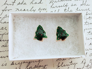 A pair of earrings made to look like sugar cookies decorated like a green Christmas tree with red sugar pearls as ornaments. The earrings are inside a white paper jewelry box.