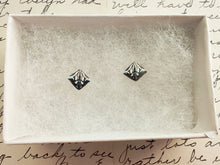 Load image into Gallery viewer, A symmetrical square abstract design of polymer clay earring studs in white, blue and metallic silver held between finger and thumb inside a white paper jewelry box.
