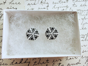 Two hexagon shaped earrings with a snowflake pattern in silver and white. The earrings are inside a white paper jewelry box.