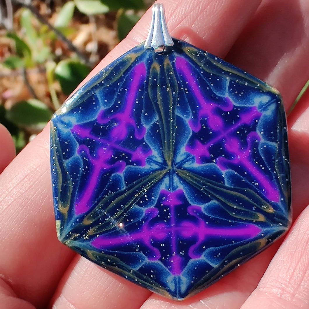 A large hexagonal pendant with a silver colored bail resting in a hand in the sunlight. The pendant is navy blue with veins of gold around the edges and radiating from the center in thirds. There are purple veins in the relative shape of a triangle throughout the pendant. 