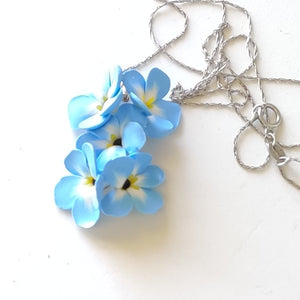 A medium pendant attached to a silver colored chain and bail. The pendant is a cluster of five Forget Me Not flowers. The flowers are pale blue and fade to white toward the center. Each petal ends in yellow at the middle and the center is black. The pendant is displayed on a white ceramic background. 