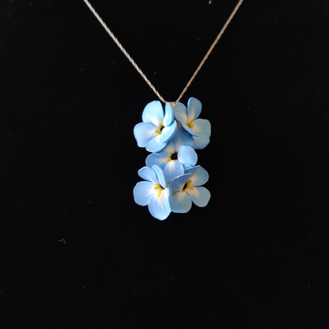 A medium pendant attached to a silver colored chain and bail. The pendant is a cluster of five Forget Me Not flowers. The flowers are pale blue and fade to white toward the center. Each petal ends in yellow at the middle and the center is black. The pendant is displayed on a black velvet background.