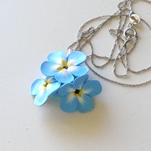 Load image into Gallery viewer, A small pendant attached to a silver colored chain and bail. The pendant is a cluster of three imitated Forget Me Not flowers. The flowers are pale blue and fade to white toward the center. Each petal ends in yellow at the middle and the center is black. The pendant is displayed on a white ceramic background. 
