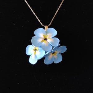A small pendant attached to a silver colored chain and bail. The pendant is a cluster of three imitated Forget Me Not flowers. The flowers are pale blue and fade to white toward the center. Each petal ends in yellow at the middle and the center is black. The pendant is displayed on a black velvet background. 