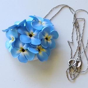 A large pendant attached to a silver colored chain displayed on a white ceramic background. The pendant is a round cluster of seven Forget Me Not imitation flowers made of polymer clay. Each of the flower petals is blue at the edges, fades to white, and ends in yellow in the middle. The center of the flowers is black. 