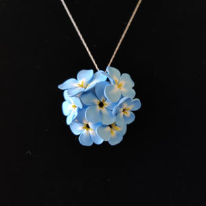 A large pendant hanging from a silver colored chain displayed on a black velvet background. The pendant is a round cluster of seven Forget Me Not imitation flowers made of polymer clay. Each of the flower petals is blue at the edges, fades to white, and ends in yellow in the middle. The center of the flowers is black. 