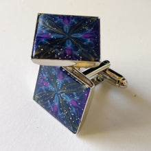 Load image into Gallery viewer, A pair of silver colored cufflinks with a square bezel on top. The bezel is filled with polymer clay with a shiny gloss finish. The clay is dark blue with veins of gold, purple, and glow in the dark clay radiating from the center. 
