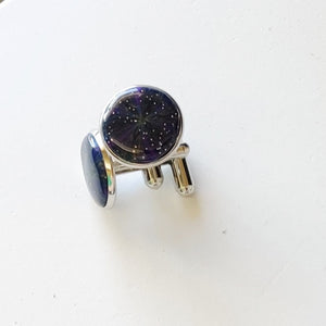 A pair of silver colored cufflinks with a bezel on top. The bezel is filled with polymer clay with a shiny gloss finish. The clay is dark blue with veins of gold, purple, and glow in the dark clay radiating from the center. 