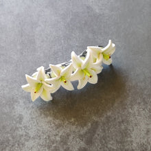 Load image into Gallery viewer, A French style hair barrette with four white lilies made of polymer clay lined up across the top. The barrette has a silver finish and four bezels which each of the lilies are set into. The hair clip is displays on a mottled dark grey surface. 
