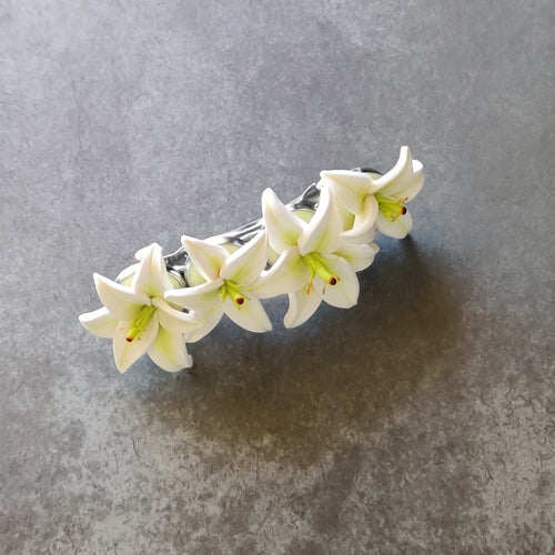 A French style hair barrette with four white lilies made of polymer clay lined up across the top. The barrette has a silver finish and four bezels which each of the lilies are set into. The hair clip is displays on a mottled dark grey surface. 