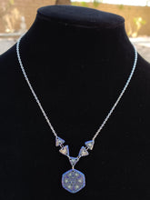 Load image into Gallery viewer, A silver colored chain neckalce connects to a series of 5 triangles in the center of a neckalce. From the center triangle hangs a hexagonal pendant. The necklace is blue, purple, and glow in the dark. 
