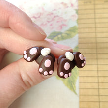 Load image into Gallery viewer, Two small bunny butt shaped earrings between finger and thumb. They are brown with pink foot pads, three toes, and a white tail.
