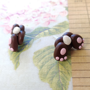 Two small bunny butt shaped earrings resting on a  flowered background. They are grey with pink food pads, three toes, and a white tail.
