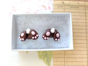 Two small bunny butt shaped earrings sitting inside a white paper jewelry box. They are brown with pink foot pads, three toes, and a white tail.