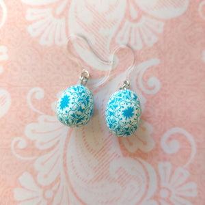 Patterned Easter egg shaped dangle earrings in a blue and white one a pink paper background.