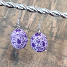 Load image into Gallery viewer, Patterned Easter egg shaped dangle earrings in a purple and white color hanging from a silver wire. 
